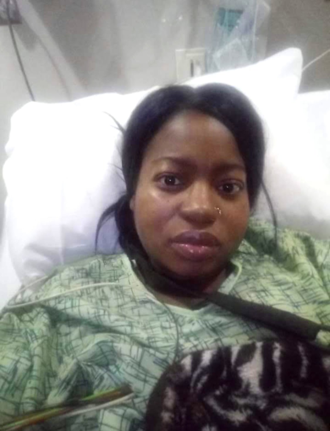 Woman lying in a hospital bed, wearing a green hospital gown, with a black and white striped blanket. Medical equipment and electrical outlets are visible behind her.