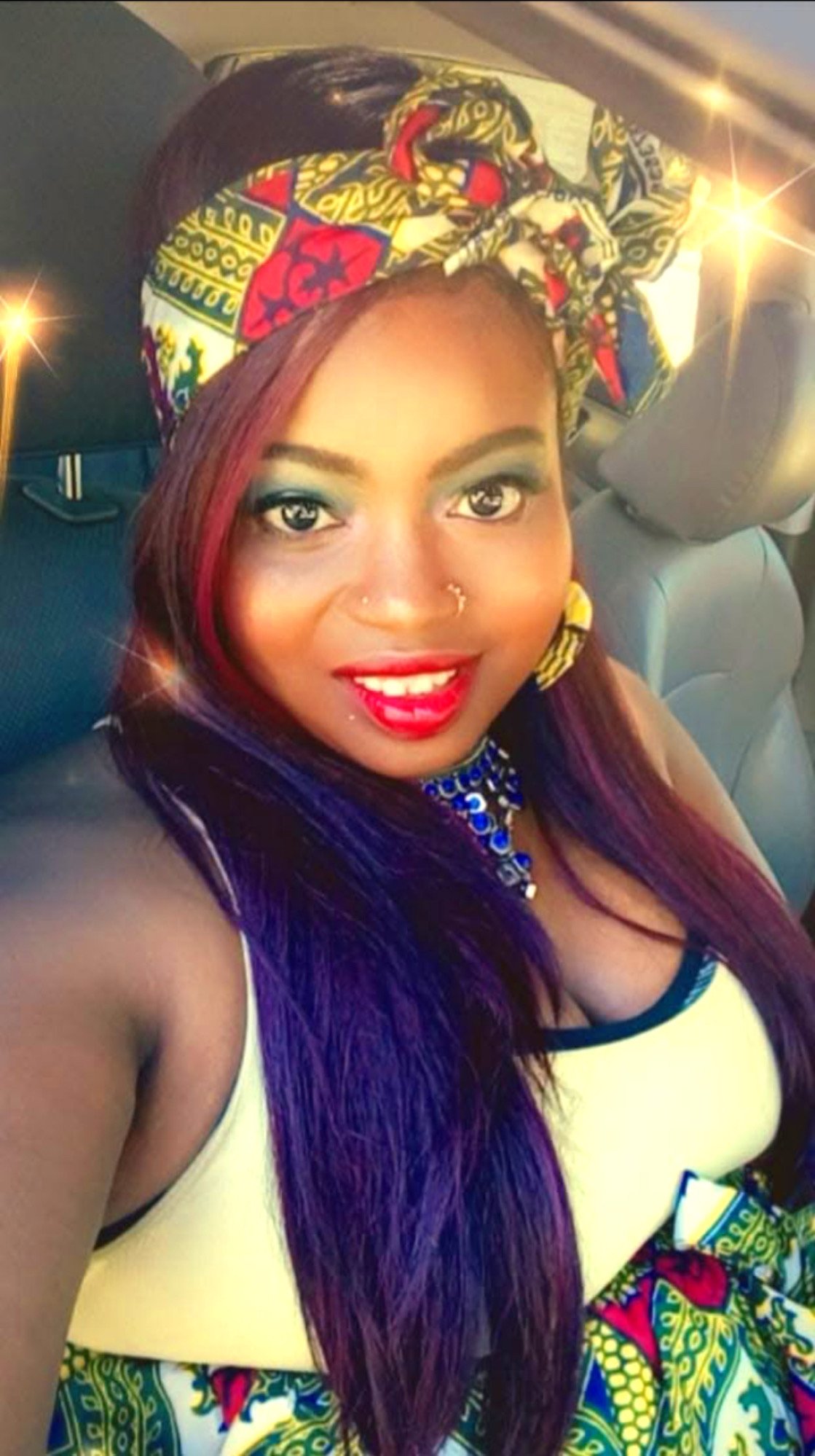 Woman smiling with colorful head wrap and dress, sitting in a car.