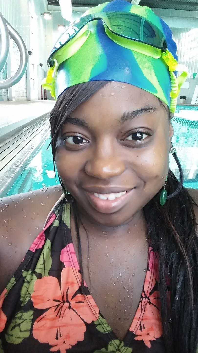 Close-up of a smiling woman at an indoor swimming pool, wearing a colorful swim cap and a floral swimsuit, with water droplets on her face and shoulders.
