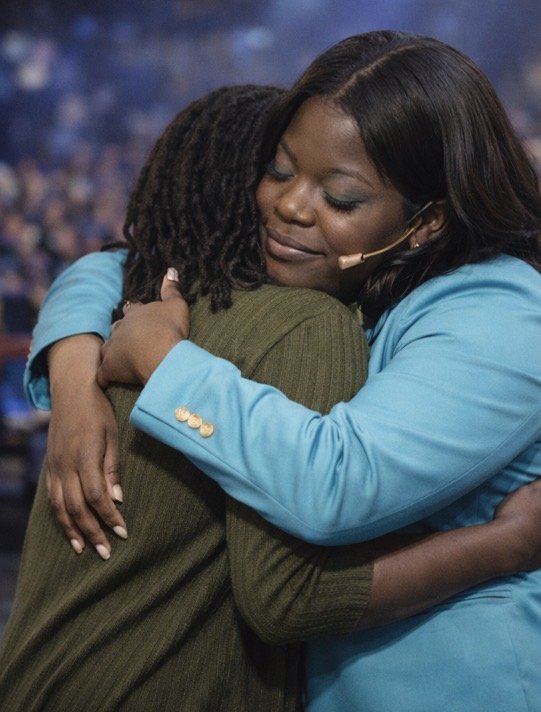 Two women hugging warmly, one with long dreadlocks and the other with straight dark hair, in an indoor setting with a blurred crowd in the background.