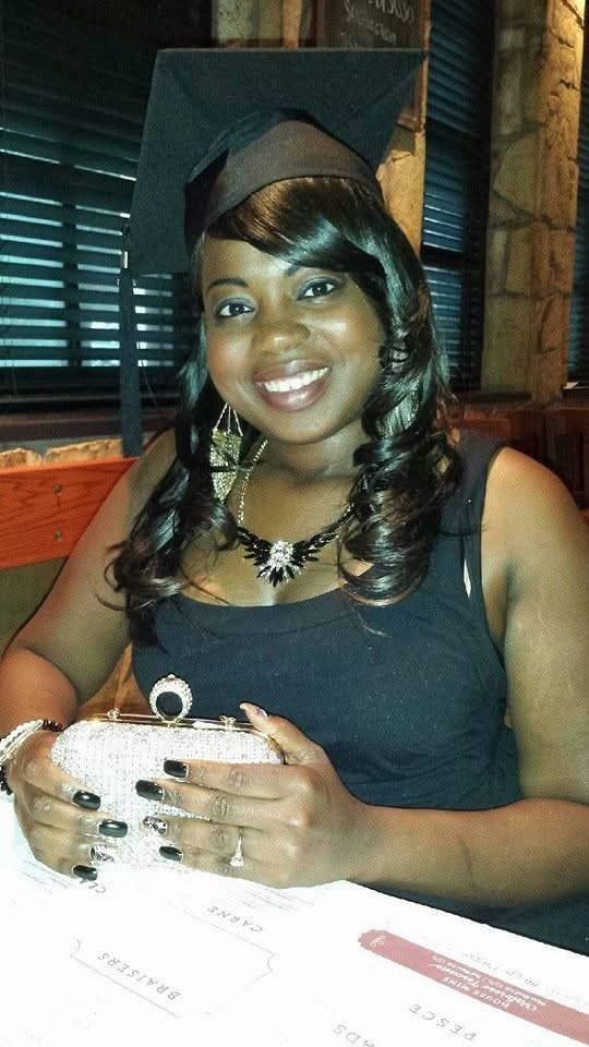 A woman in a black dress and a graduation cap, sitting at a table in a restaurant or cafe, holding a small sparkling clutch purse and smiling at the camera.