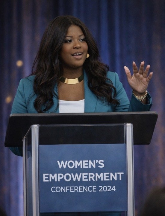 A woman speaking at a conference behind a podium with a sign that says 'Women's Empowerment Conference 2024'. She is gesturing with one hand and wearing a teal blazer and a gold necklace.