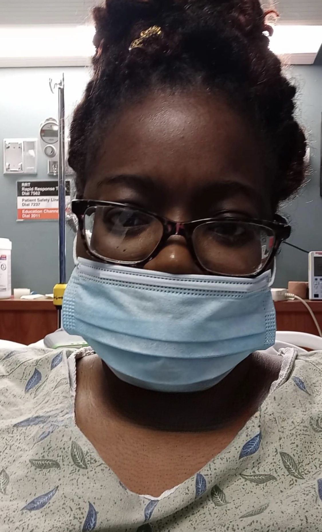 A woman wearing glasses and a face mask sitting in a hospital room or medical facility.