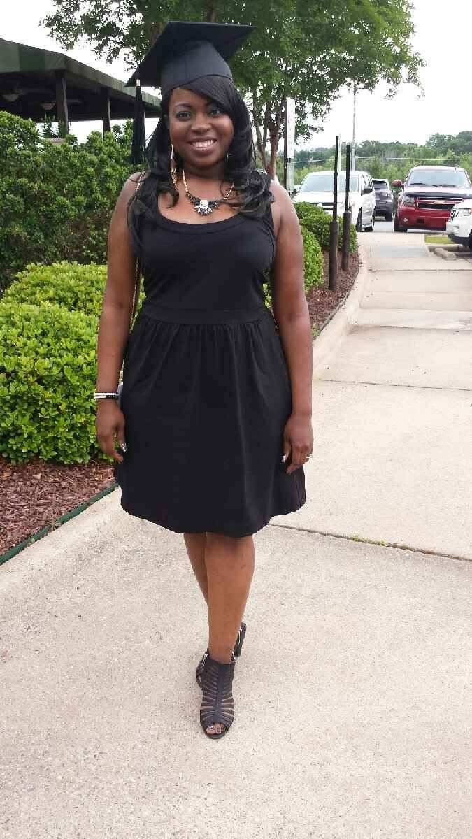 A young woman with dark hair wearing a black graduation cap and dress, standing on a sidewalk with cars and greenery in the background, smiling at the camera.