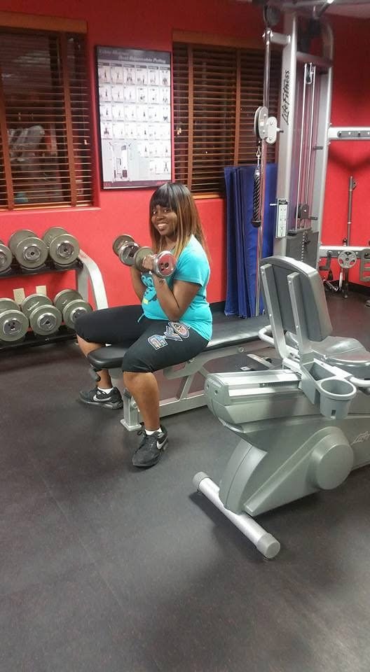 A woman is sitting on a gym bench lifting a dumbbell in a fitness room. There are weight racks, exercise equipment, and a red wall with windows in the background.