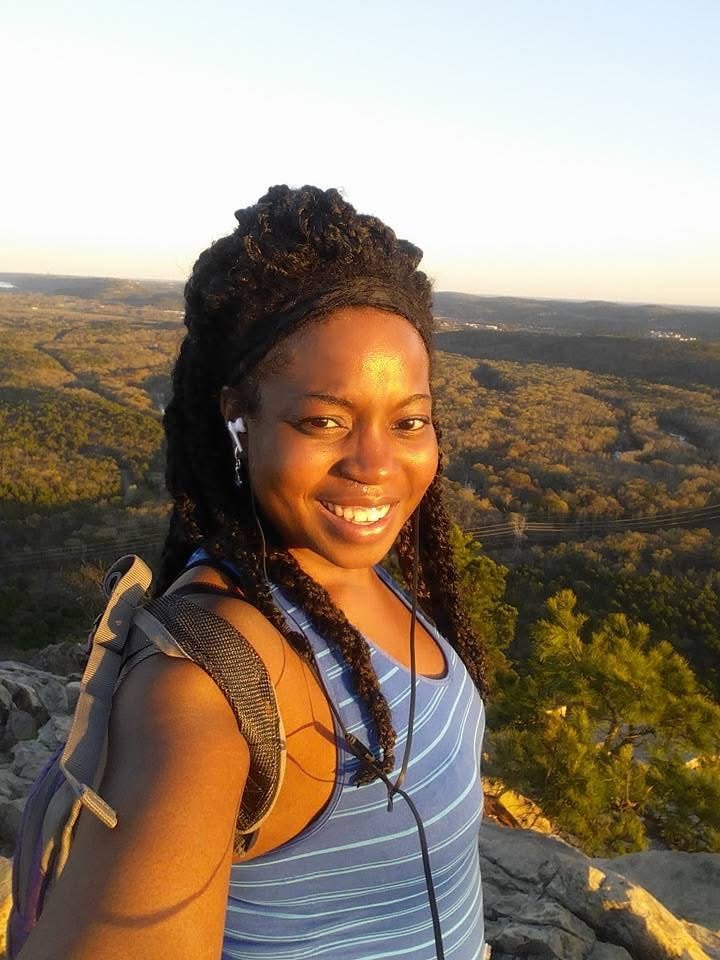 A woman smiling outdoors during sunset, wearing a striped tank top, with a backpack and earbuds, with a scenic landscape of rolling hills and trees in the background.