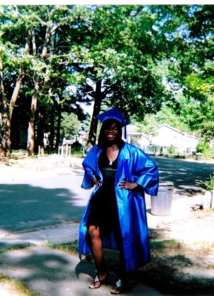 A young woman in a blue graduation gown and cap standing outdoors on a street with trees and houses in the background.