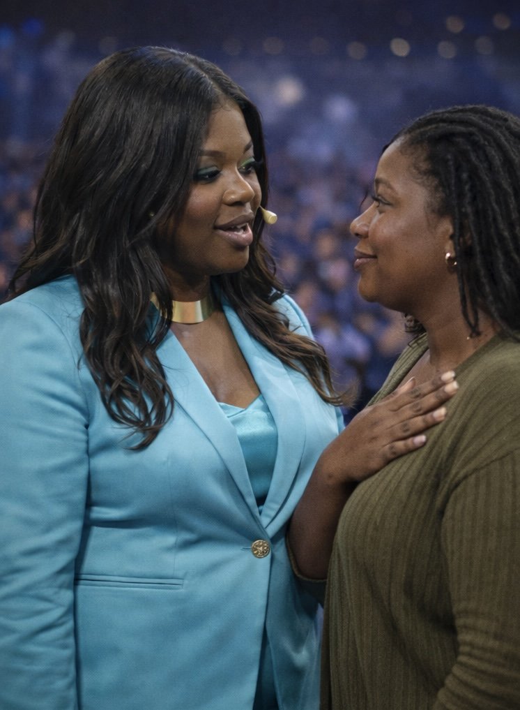 Two women, one in a blue blazer and the other in a green sweater, face each other in an emotional moment, one touching the other's chest in a crowded indoor setting.