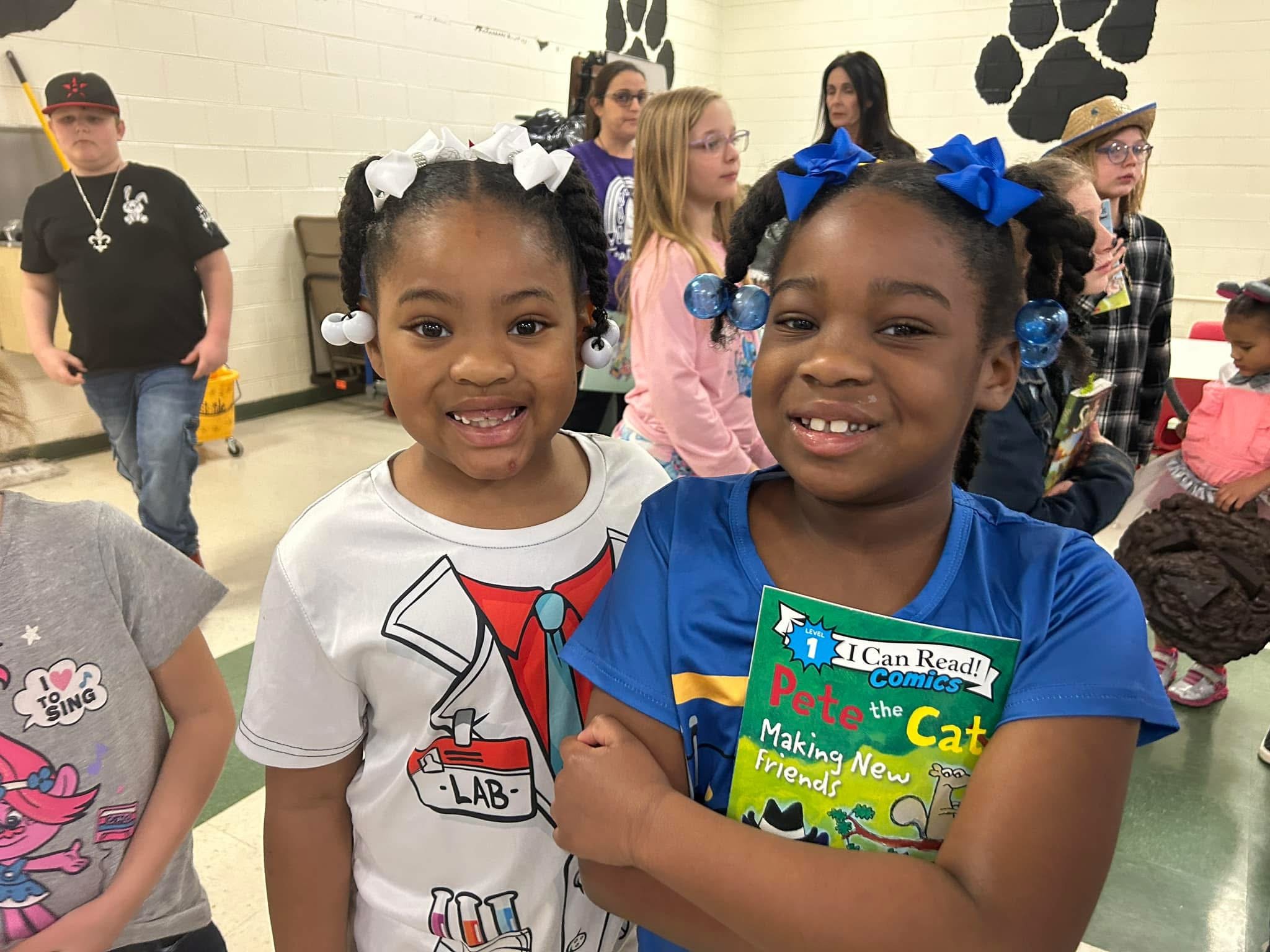 Two young girls smiling at the camera, one holding a comic book titled "Pete the Cat ». They are in a room with other children and adults in the background.
