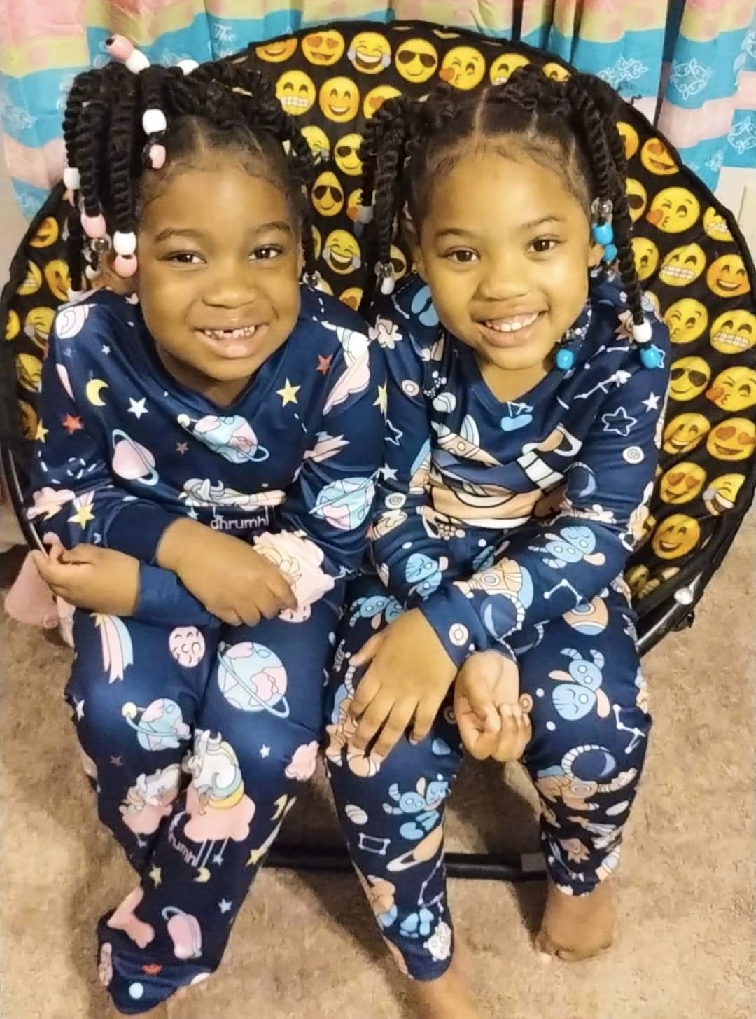Two young girls with braided hair adorned with beads, smiling while sitting together on a chair with a black cover decorated with yellow emojis, wearing matching space-themed pajamas with planets, stars, and rockets.