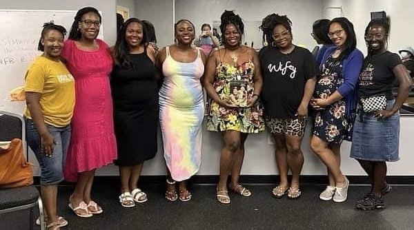 Group of eight women standing together indoors, smiling, with a wall and mirror behind them.