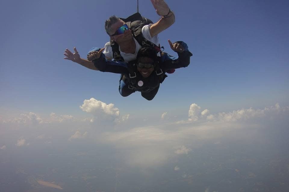 A man and woman smiling and giving thumbs-up during a tandem skydiving jump, freefalling against a background of blue sky and clouds.