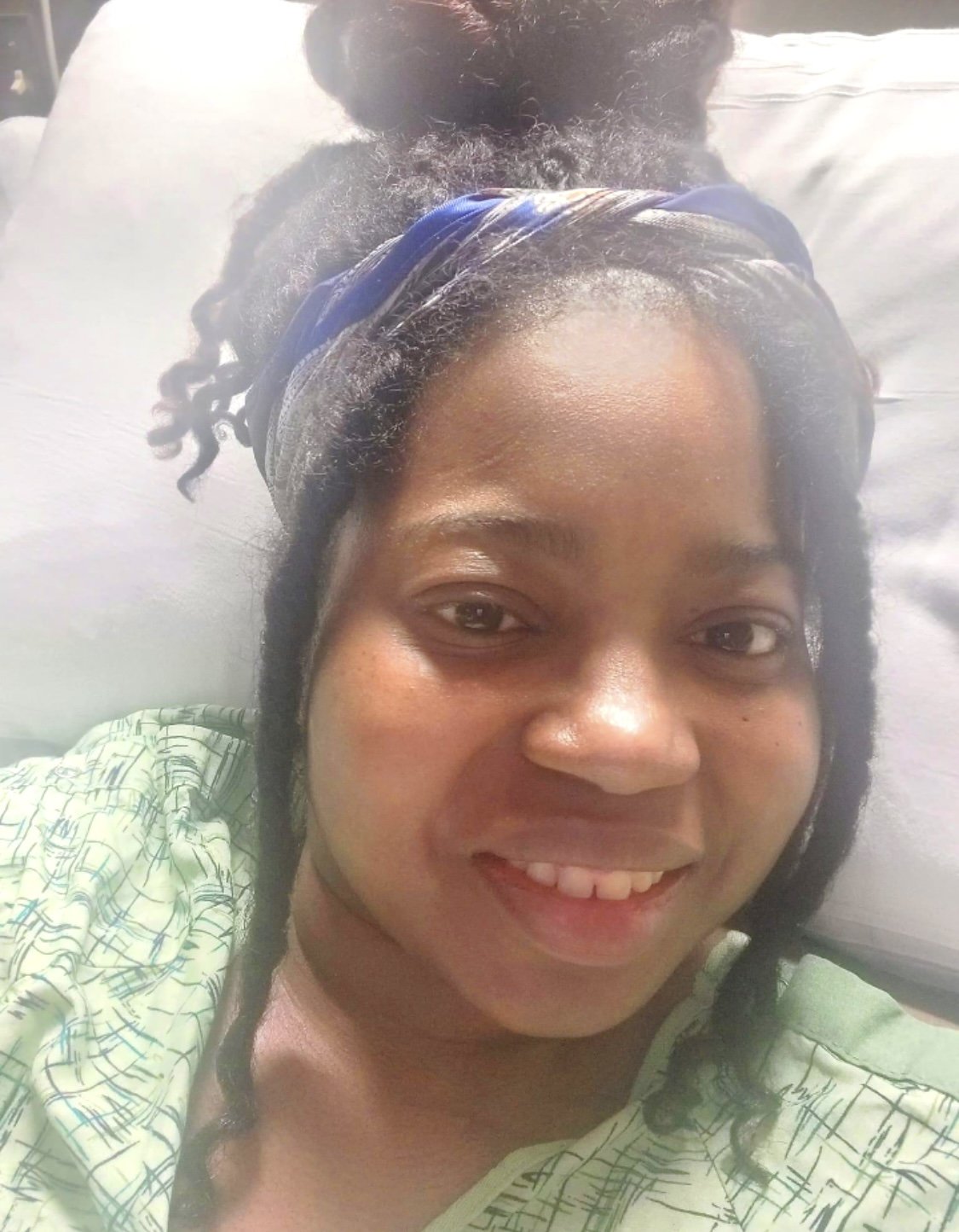 A woman with dark hair styled in braids and a headscarf lying in a hospital bed, smiling at the camera.