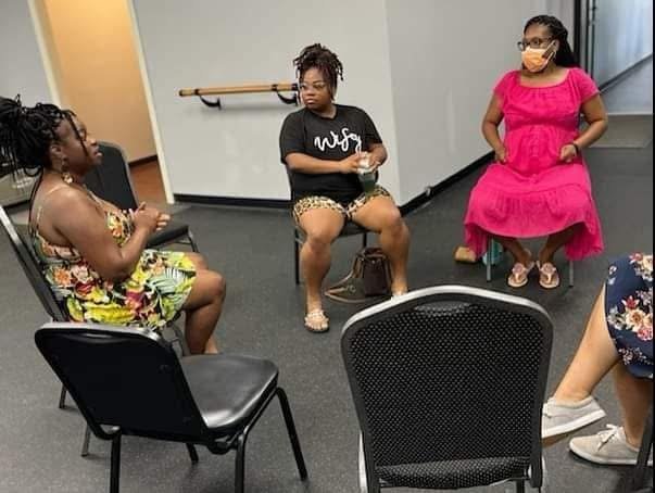 Three women sitting in a circle in a room. One woman is wearing a floral dress, another is in a black shirt and shorts, and the third woman is in a pink dress and mask. They are engaged in conversation.
