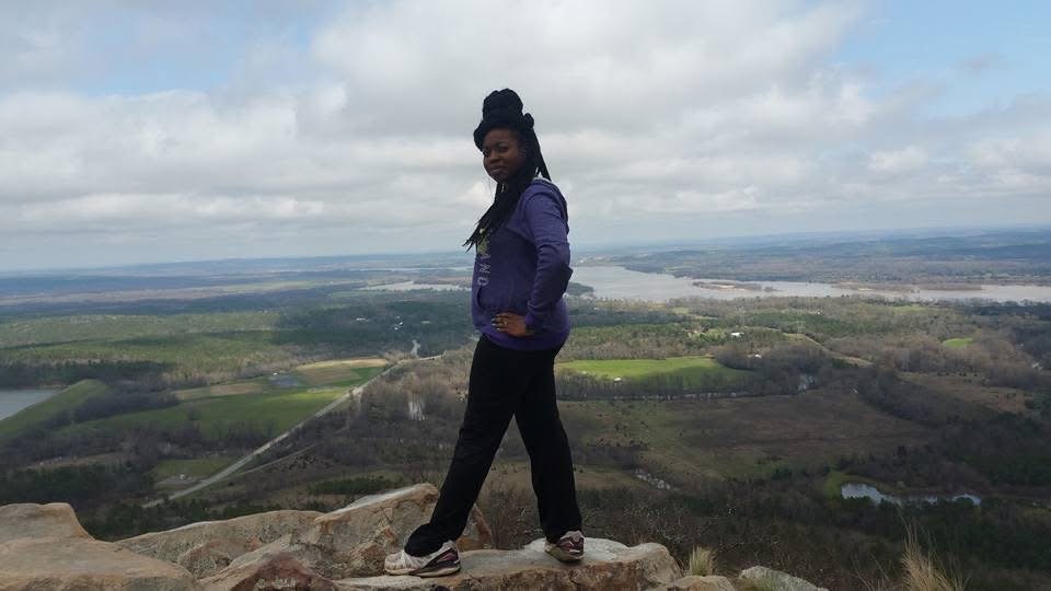 A woman standing on a rocky cliff overlooking a river and expansive landscape with trees and fields, under a cloudy sky.