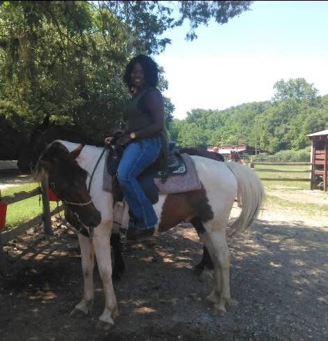 A woman riding a small horse or pony outdoors in a rural setting with trees and farm buildings in the background.