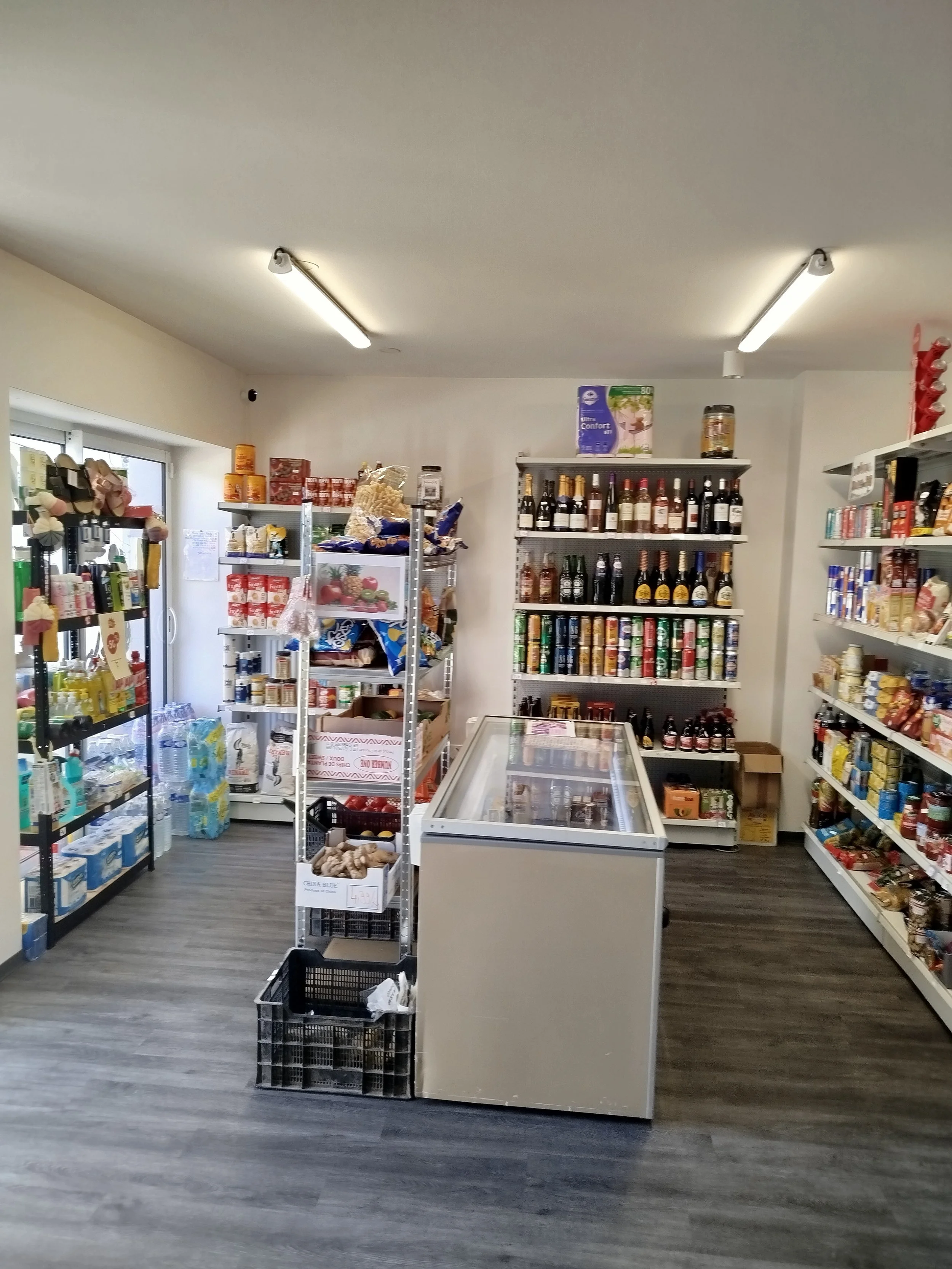 Intérieur d'une petite épicerie avec des étagères remplies de produits alimentaires, un congélateur en centre et un sol en bois gris.