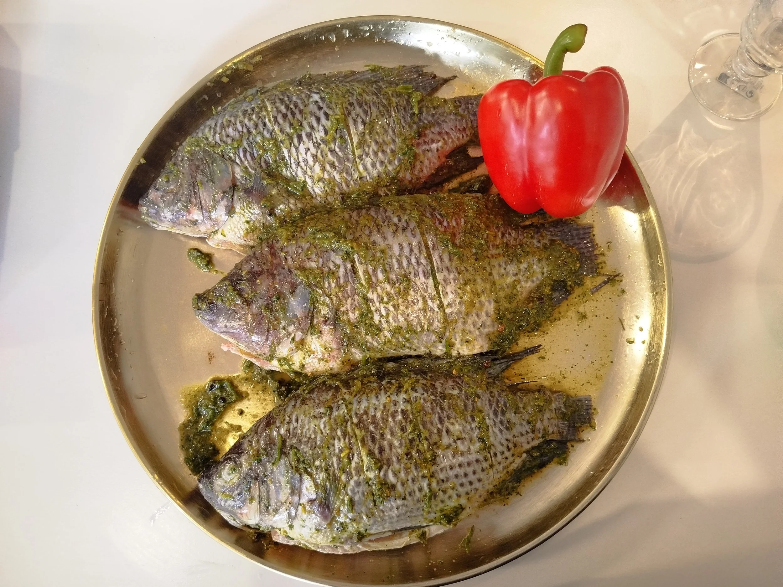 Trois poissons assaisonnés avec une sauce verte, accompagnés d'un poivron rouge sur un plateau en métal.