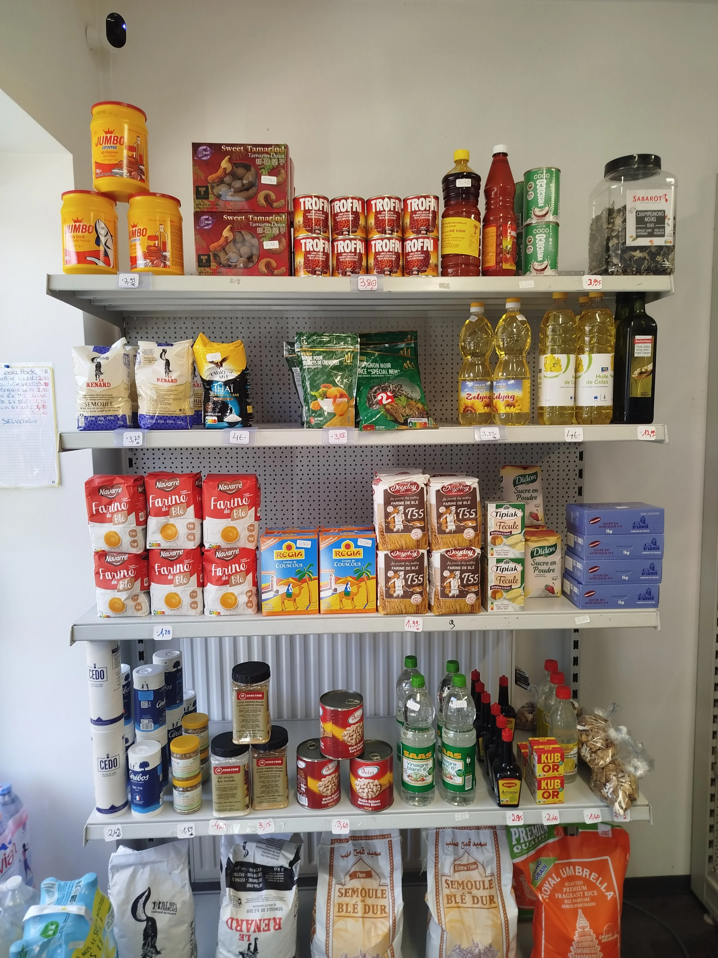Rayonnage avec produits alimentaires tels que farines, huiles, biscuits, épices, conserves, et boissons en bouteilles.