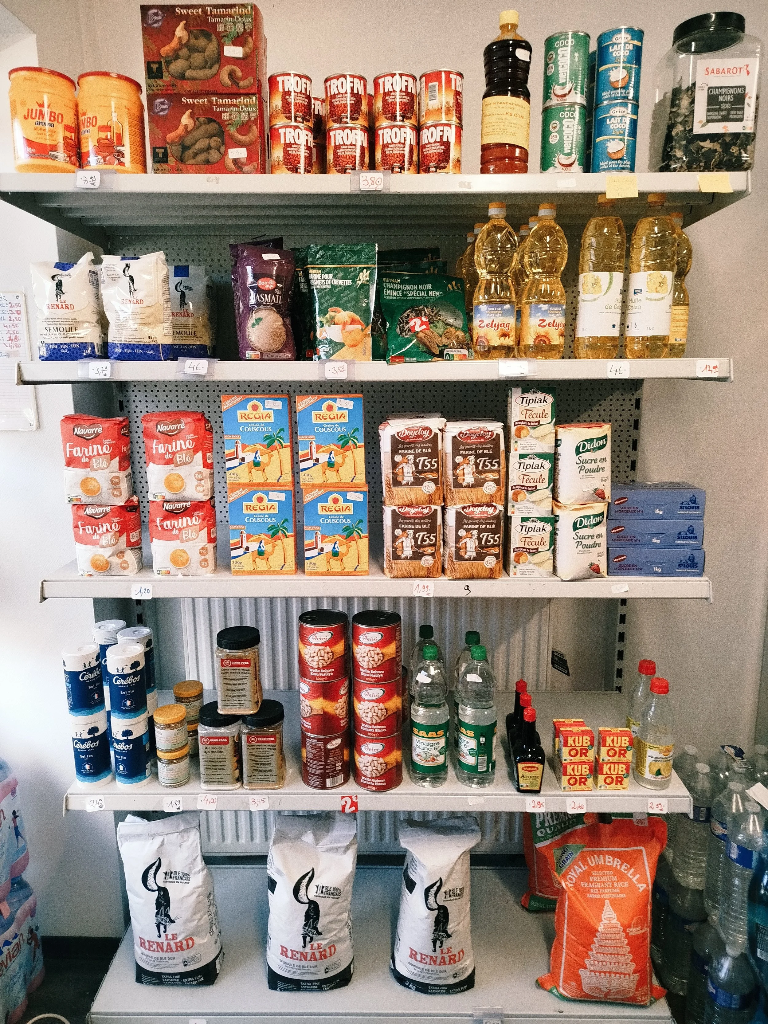 Étagère de supermarché avec divers produits alimentaires, notamment des conserves, huiles, farines, riz, et bouteilles d'eau.