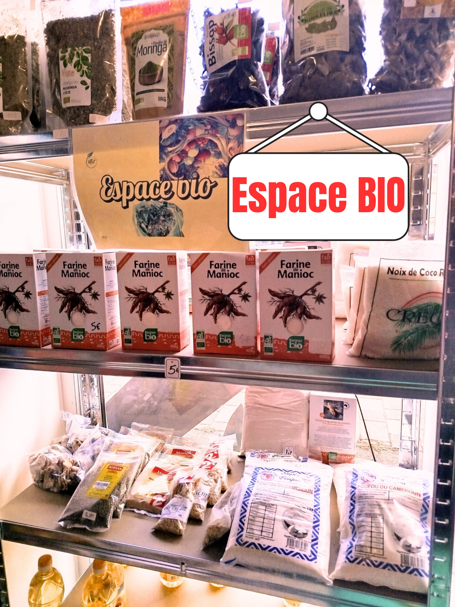 Une étagère en magasin avec des produits biologiques, notamment des sachets de farine de manioc, des noix de coco, et d'autres aliments. Un panneau indique 'Espace BIO'.