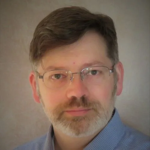 A middle-aged man with glasses and a beard, wearing a blue collared shirt, looking at the camera against a neutral background.