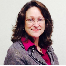 A woman with shoulder-length brown hair and glasses, wearing a gray blazer and pink blouse, smiling
