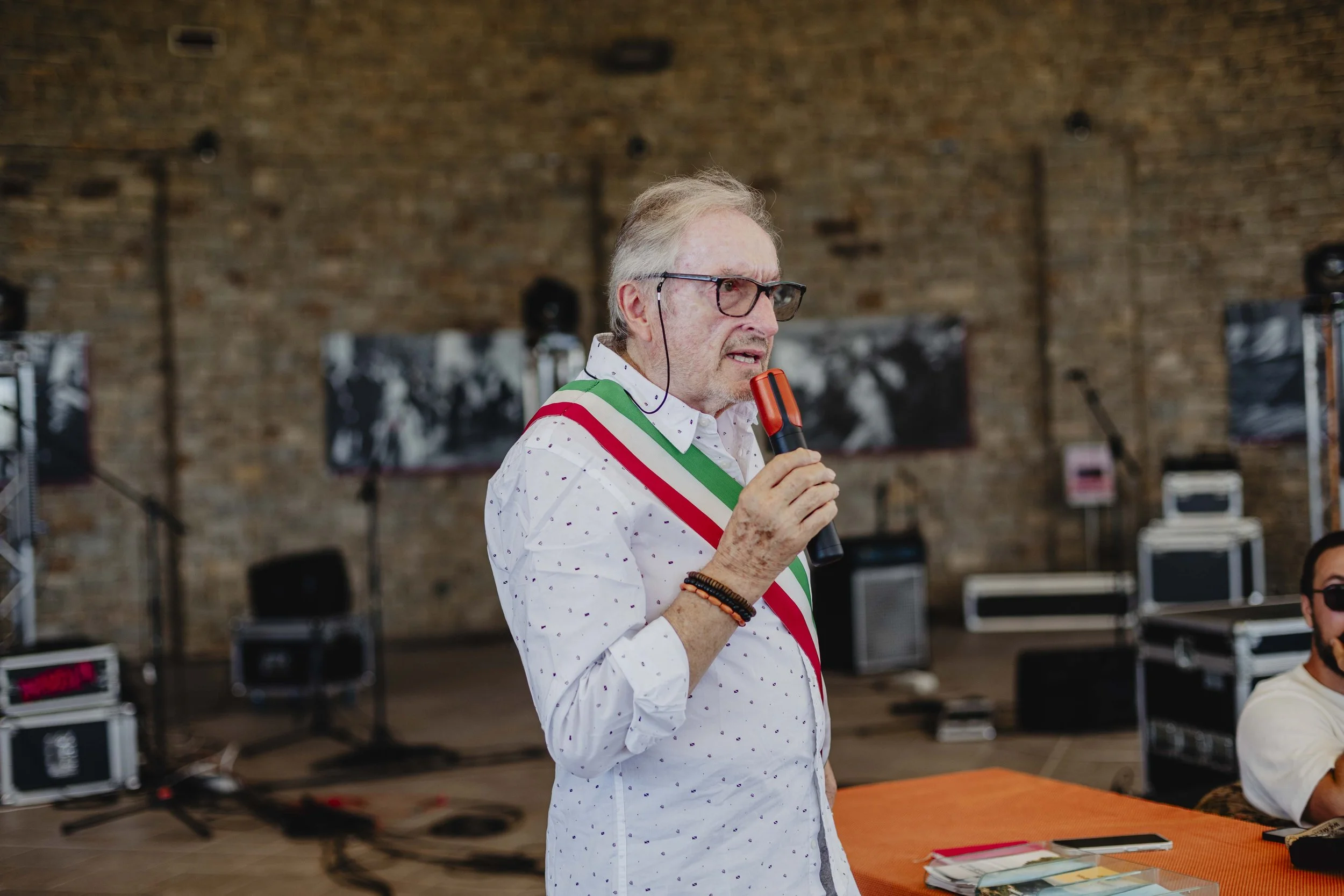 Uomo anziano con occhiali e maglietta bianca con puntini colorati parla al microfono durante un evento, con sfondo di parete in mattoni e apparecchiature audio.