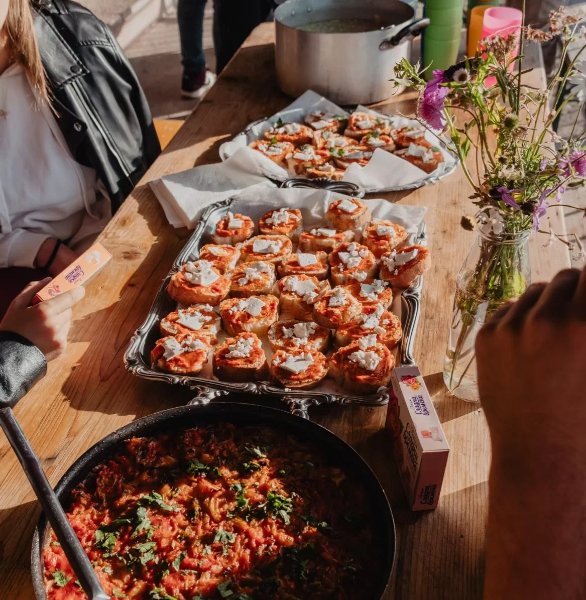 Un tavolo di legno con piatti di antipasti, tra cui bruschette con formaggio e pomodoro, un piatto di sugo di pomodoro con erbe e una brocca di acqua, decorato con un mazzo di fiori.