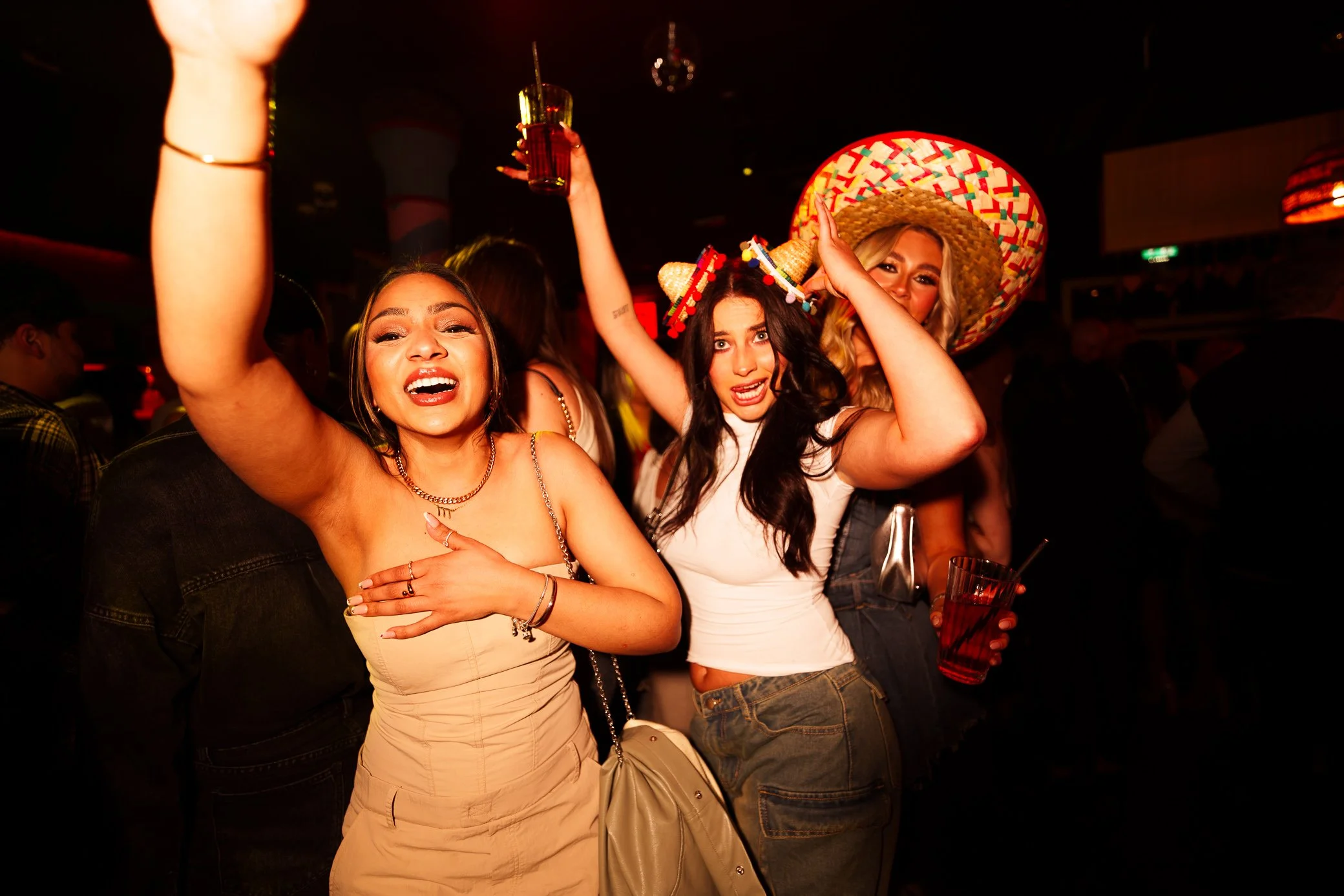 Three young women celebrating at a party, holding drinks, with one wearing a large colorful sombrero, in a lively, dark bar or club setting.
