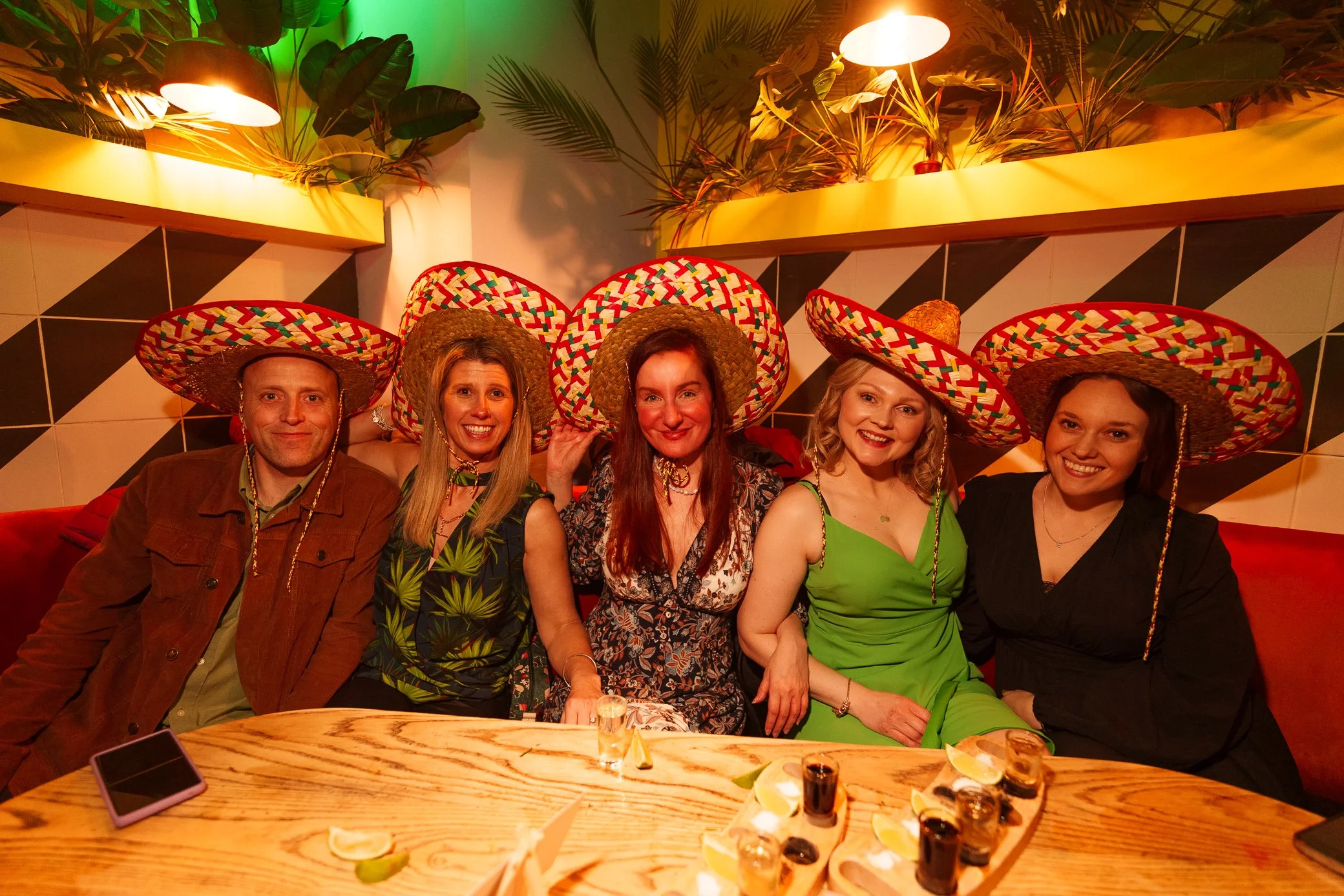 Five people sitting at a wooden table wearing colorful sombreros in a festive setting with green plants and colorful lighting.