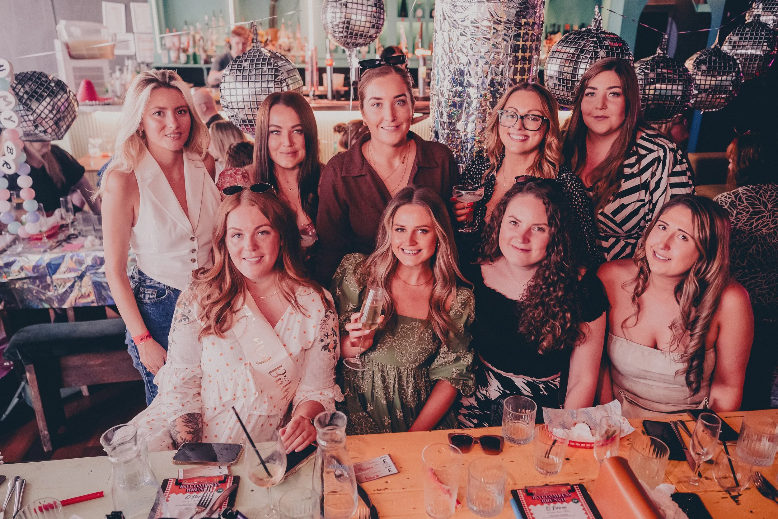 A group of women celebrating at a party, with some wearing party hats and sashes, in a decorated venue with disco balls and festive decor.