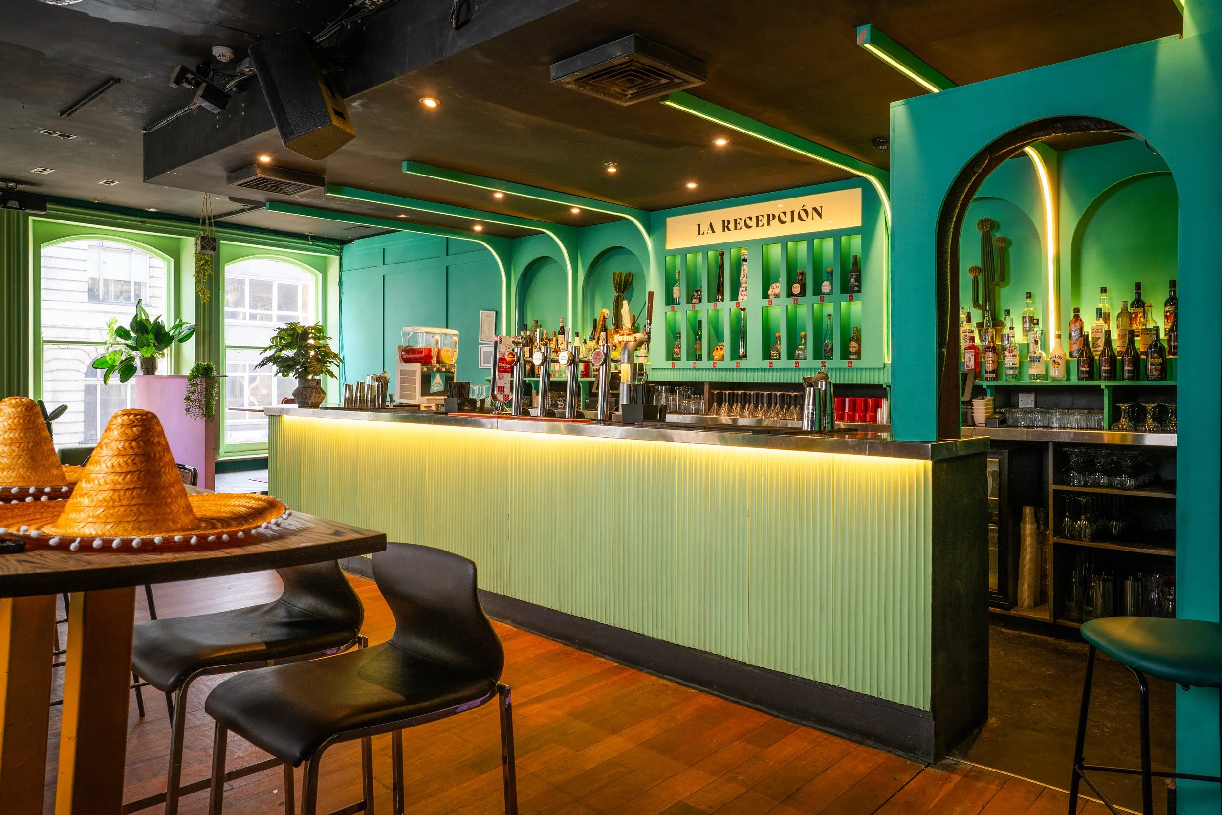 Colorful bar with yellow illuminated counter, turquoise and green walls, and Mexican sombreros on a table in foreground.