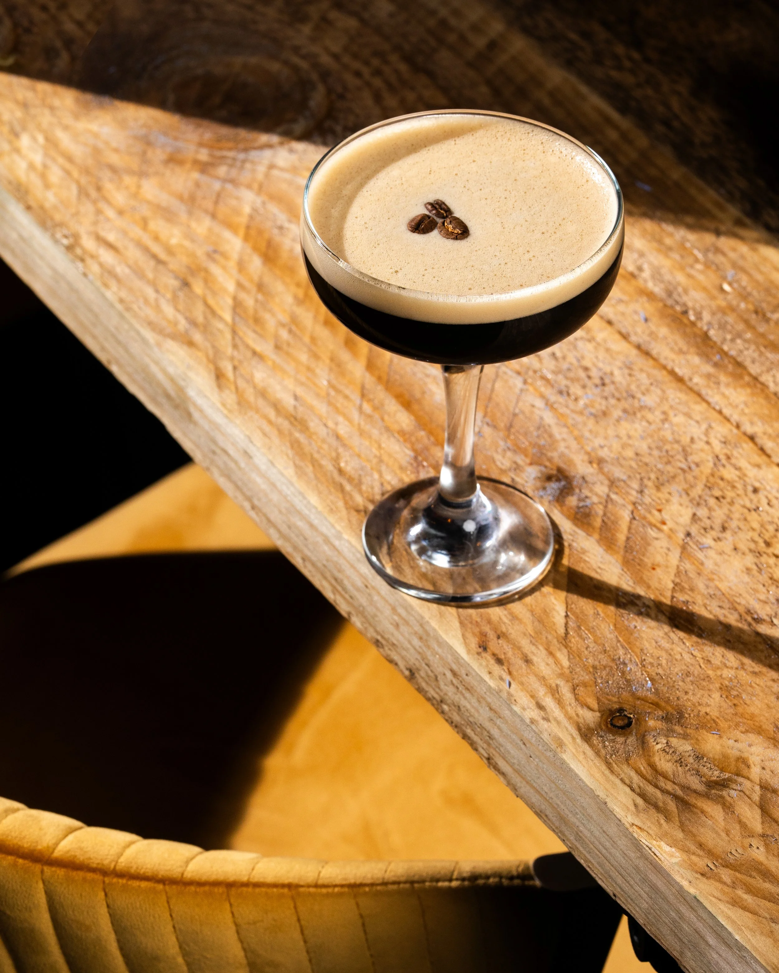A cocktail glass with dark drink topped with frothy cream and coffee beans, placed on a rustic wooden table.