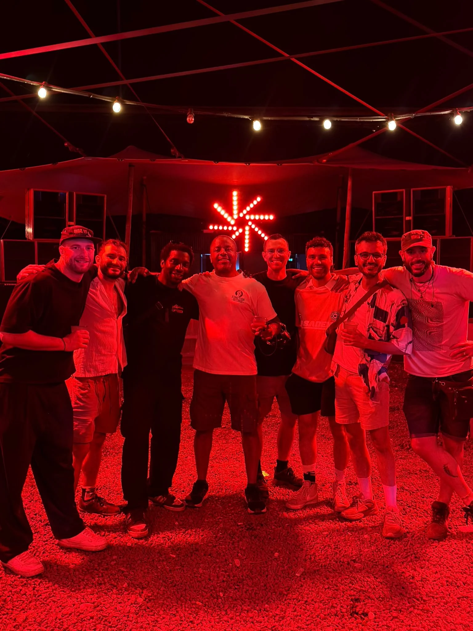 Group of eight young men standing indoors under red lighting, with a red neon sign in the shape of an asterisk behind them, wearing casual clothes and smiling.