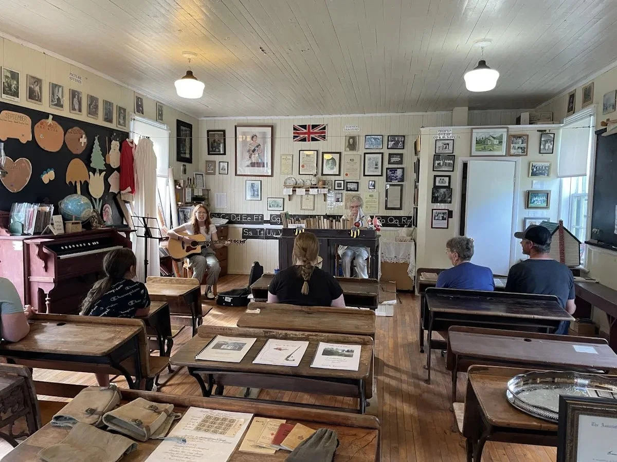 A small classroom with a few students and two performers, one playing the guitar and the other at a piano, while the students sit at desks facing the performers. The classroom has framed pictures and decorations on the walls, and a blackboard with ed