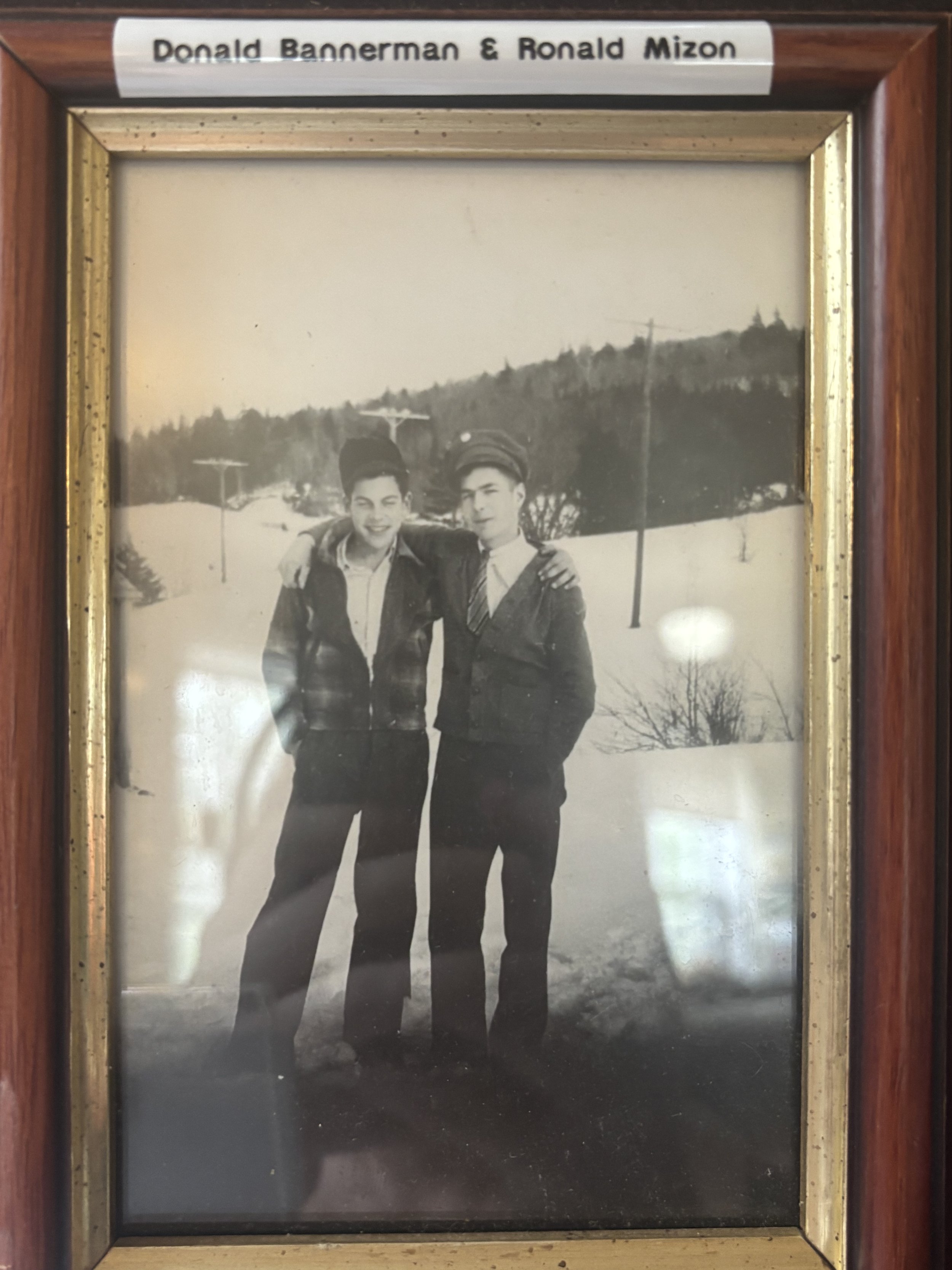 Black and white photo of two young men standing outdoors in winter, smiling and with arms around each other, with snow-covered ground and hills in the background.