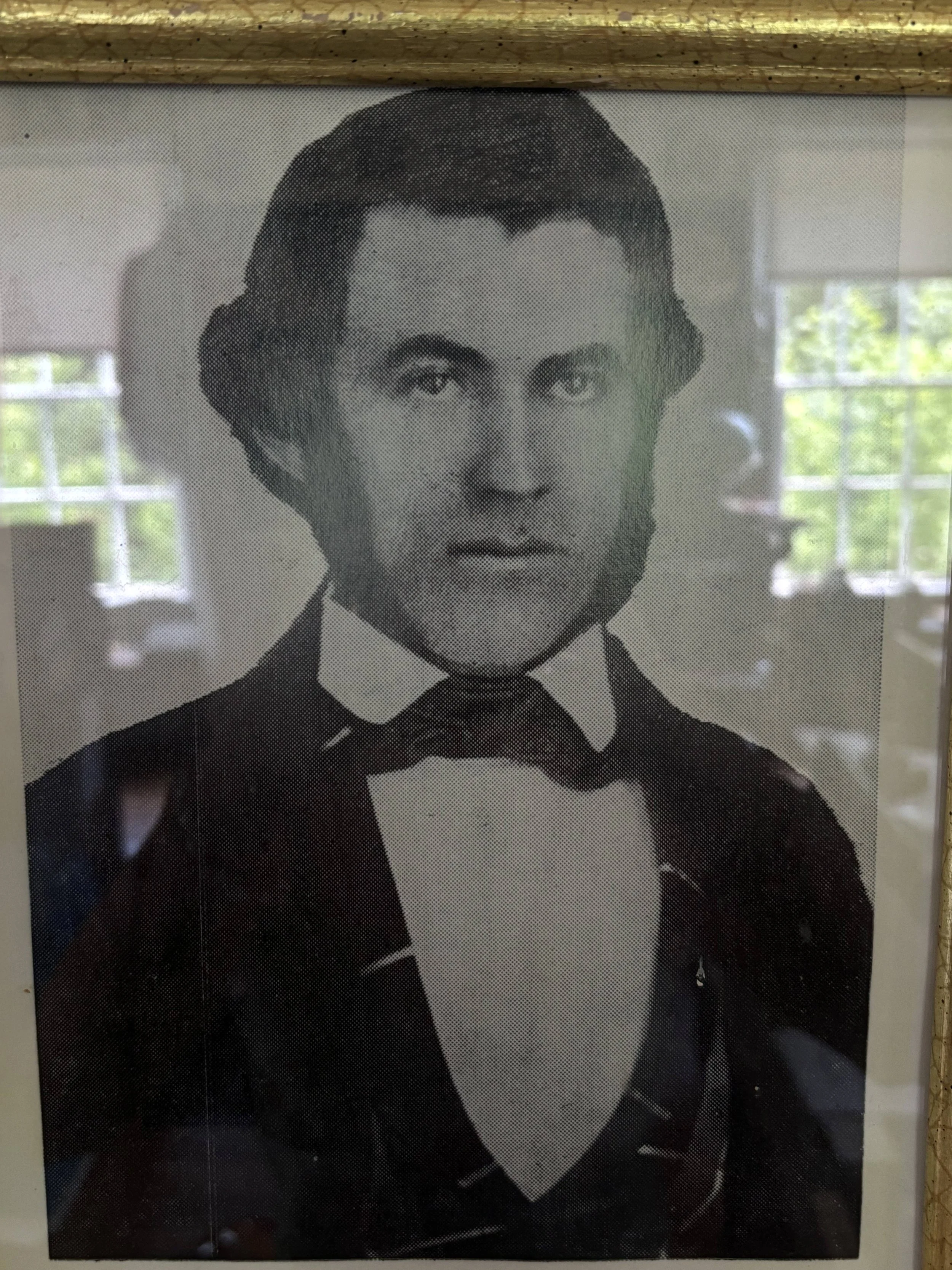 Black and white portrait of a man in a 19th-century style tuxedo with a bow tie and a white shirt, with reflections of windows and green trees in the background.
