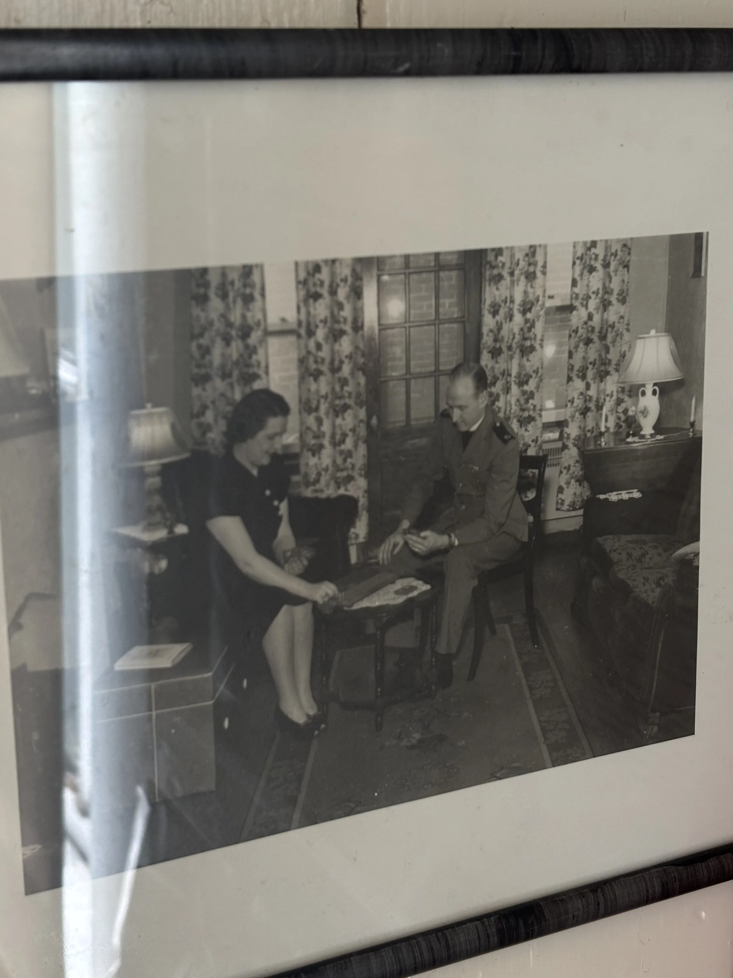 Black and white photograph of a woman and a man sitting in a living room, engaged in a conversation, with the woman holding an object and the man reading a magazine or book. The room has floral curtains, a lamp, and a sofa.