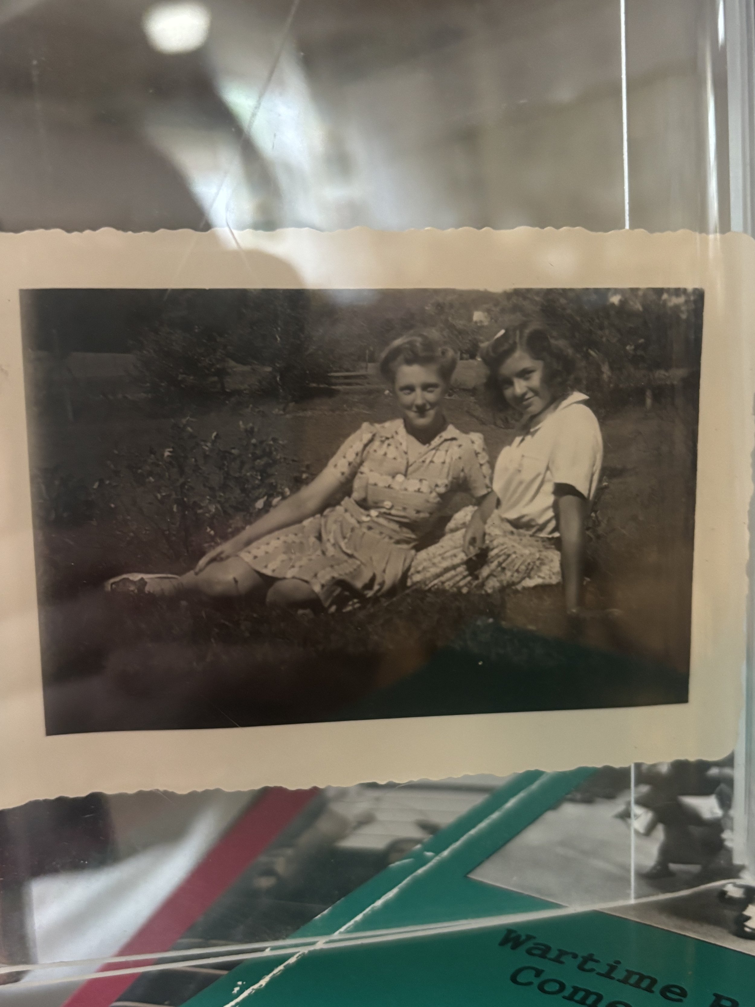 Black and white photograph of two women sitting and lying on the grass outdoors, smiling at the camera, with trees in the background.
