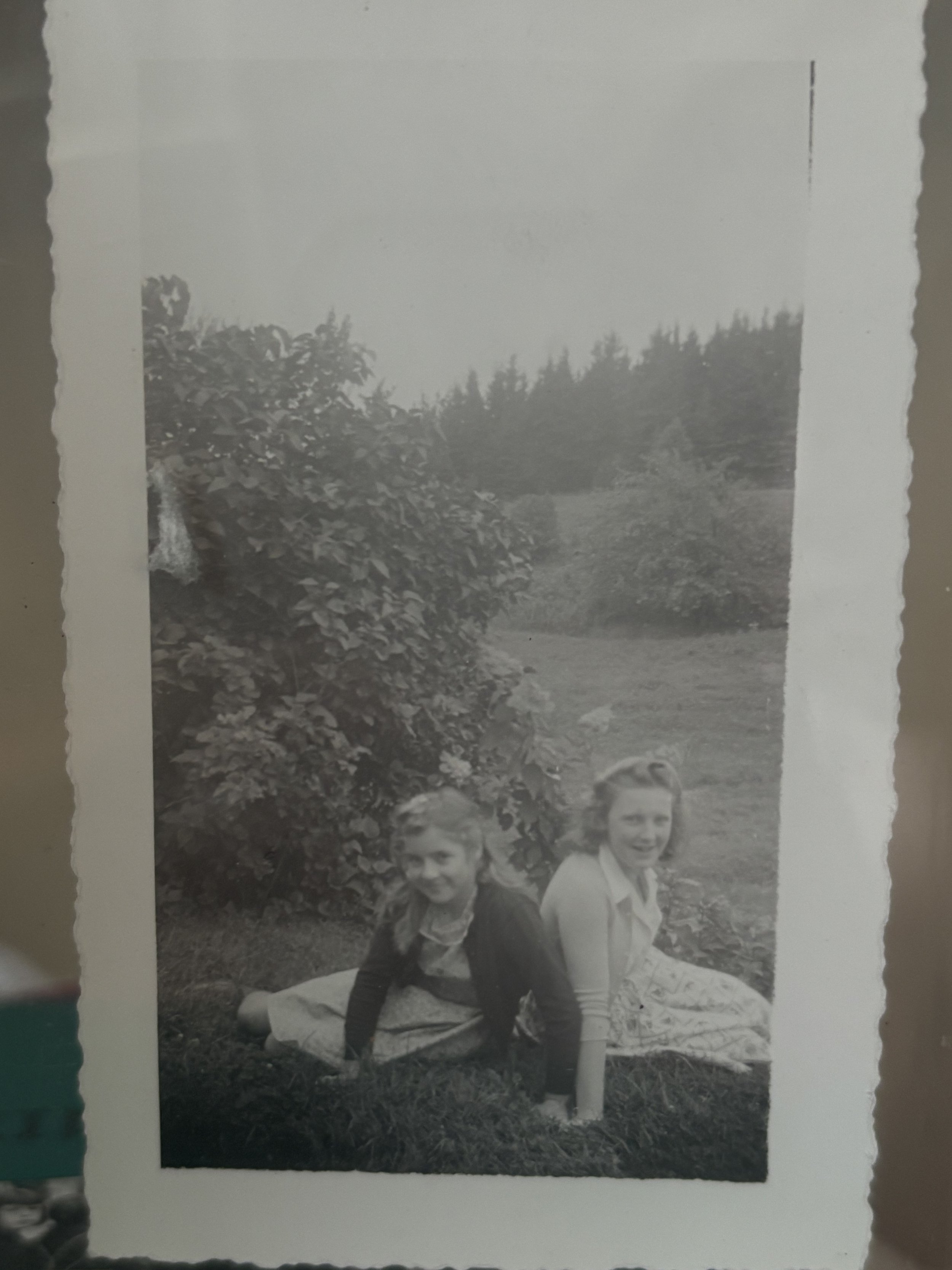 Two young girls sitting on grass in an outdoor setting, smiling at the camera, with trees and bushes in the background.
