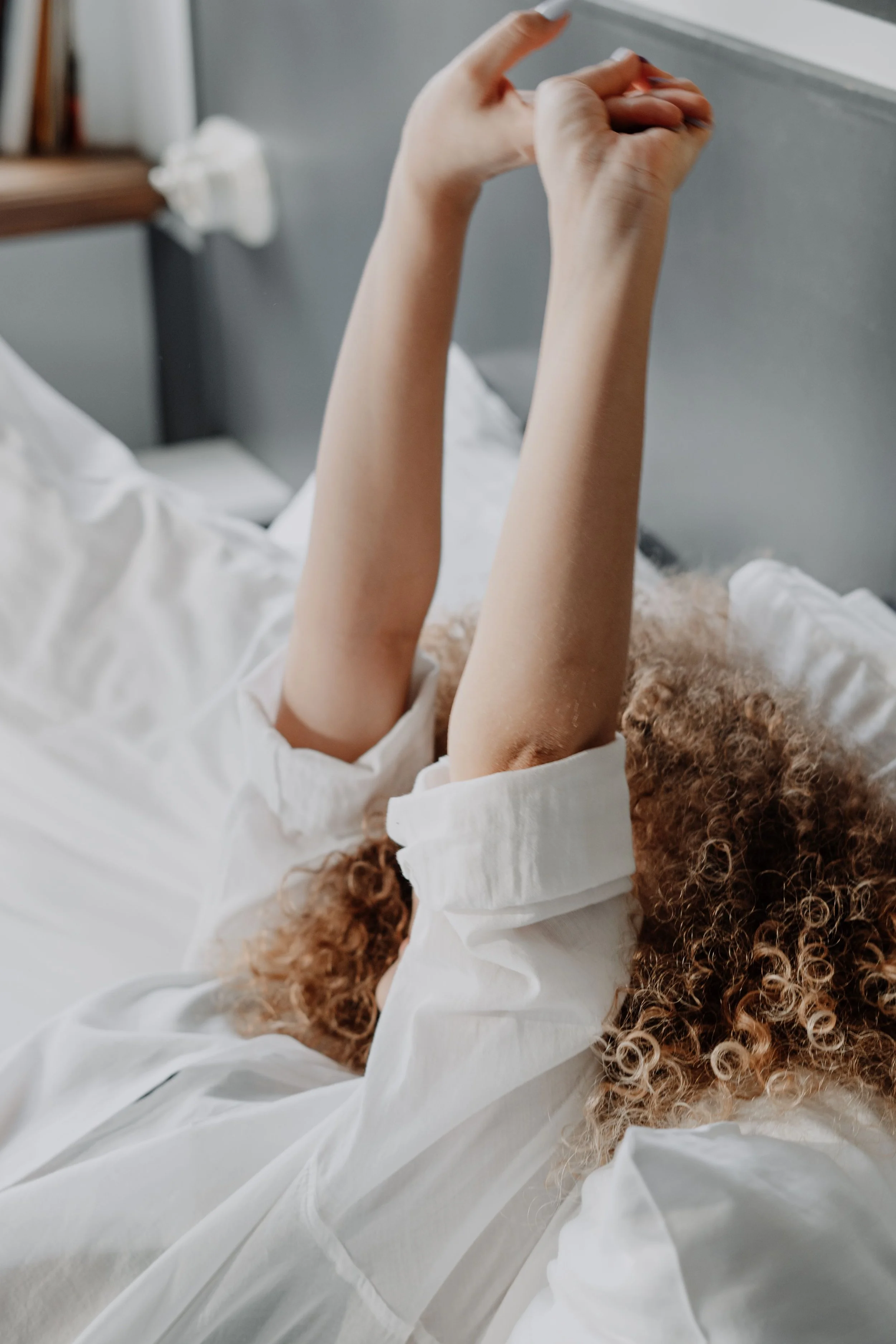 A person lying in bed with arms raised above their head, wearing a white shirt, with a curly-haired dog resting on their chest. The bed has white sheets, and a bookshelf is visible in the background.