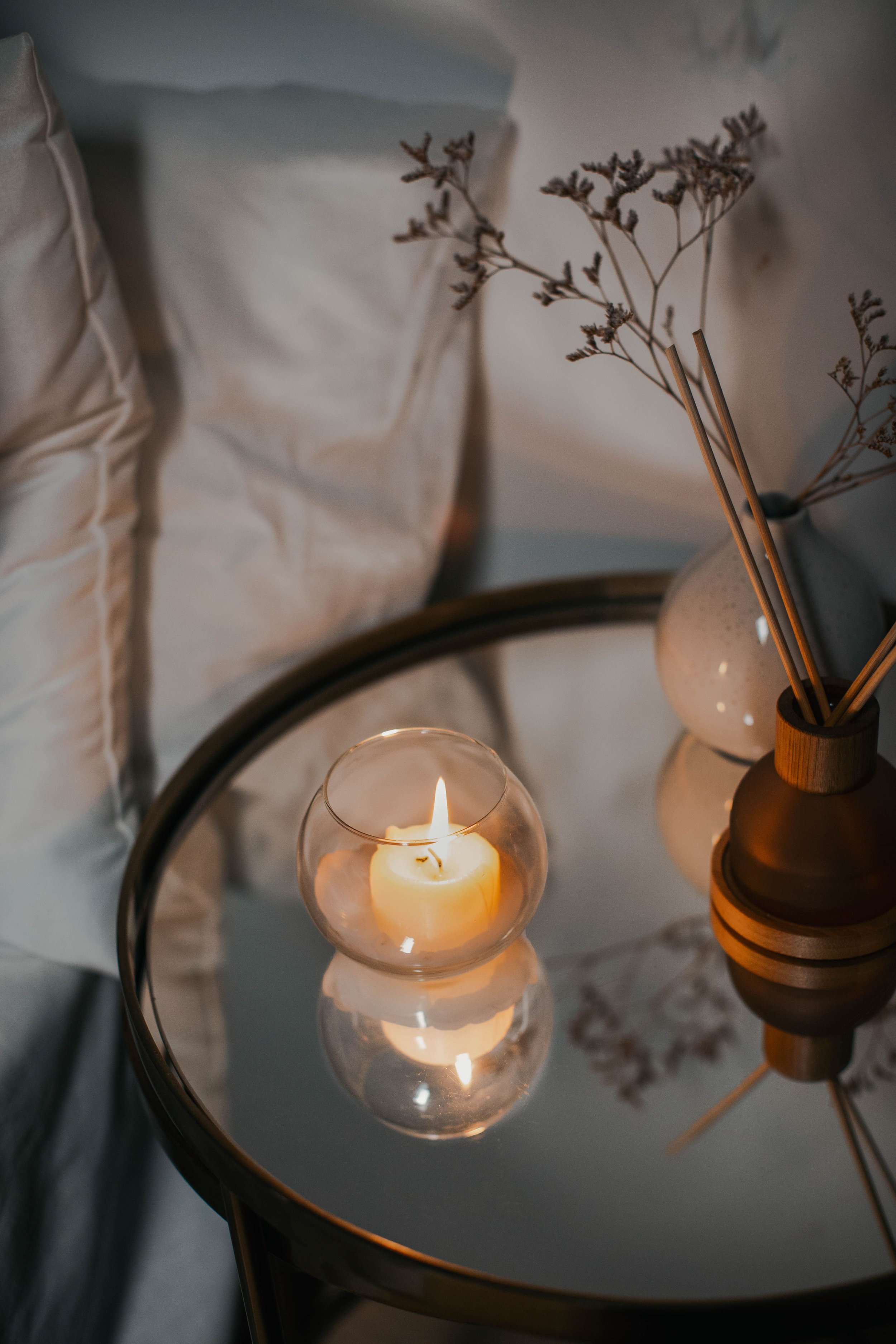 A lit candle inside a glass holder reflects in the surface of a black, round mirror table. Next to the candle, there is a brown ceramic or wooden vase holding dried, brownish flowers or branches. Behind the table, there are light-colored pillows on a bed or sofa.