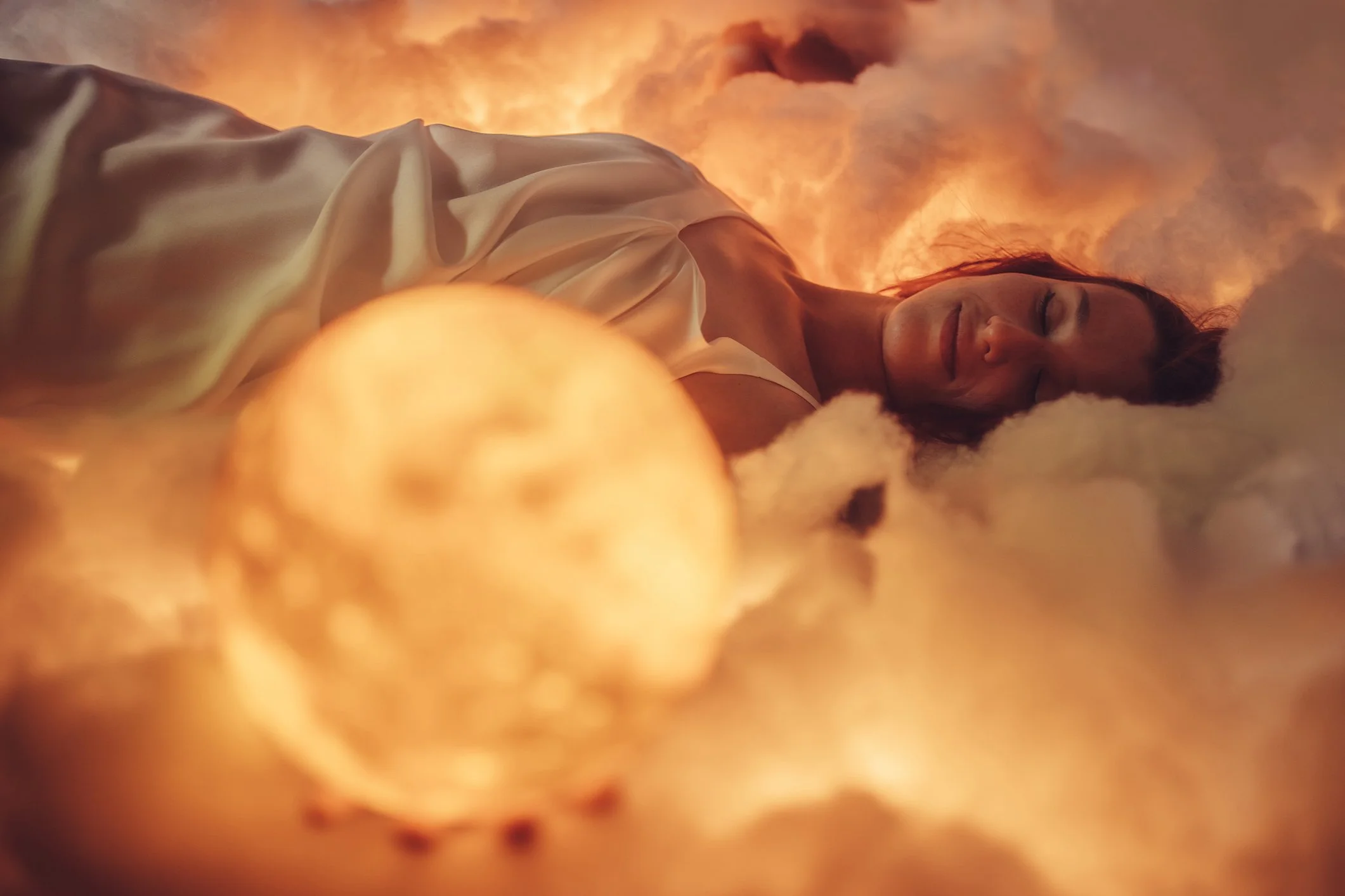 A woman lying on her side with her eyes closed, surrounded by glowing clouds of orange and yellow smoke or fire.