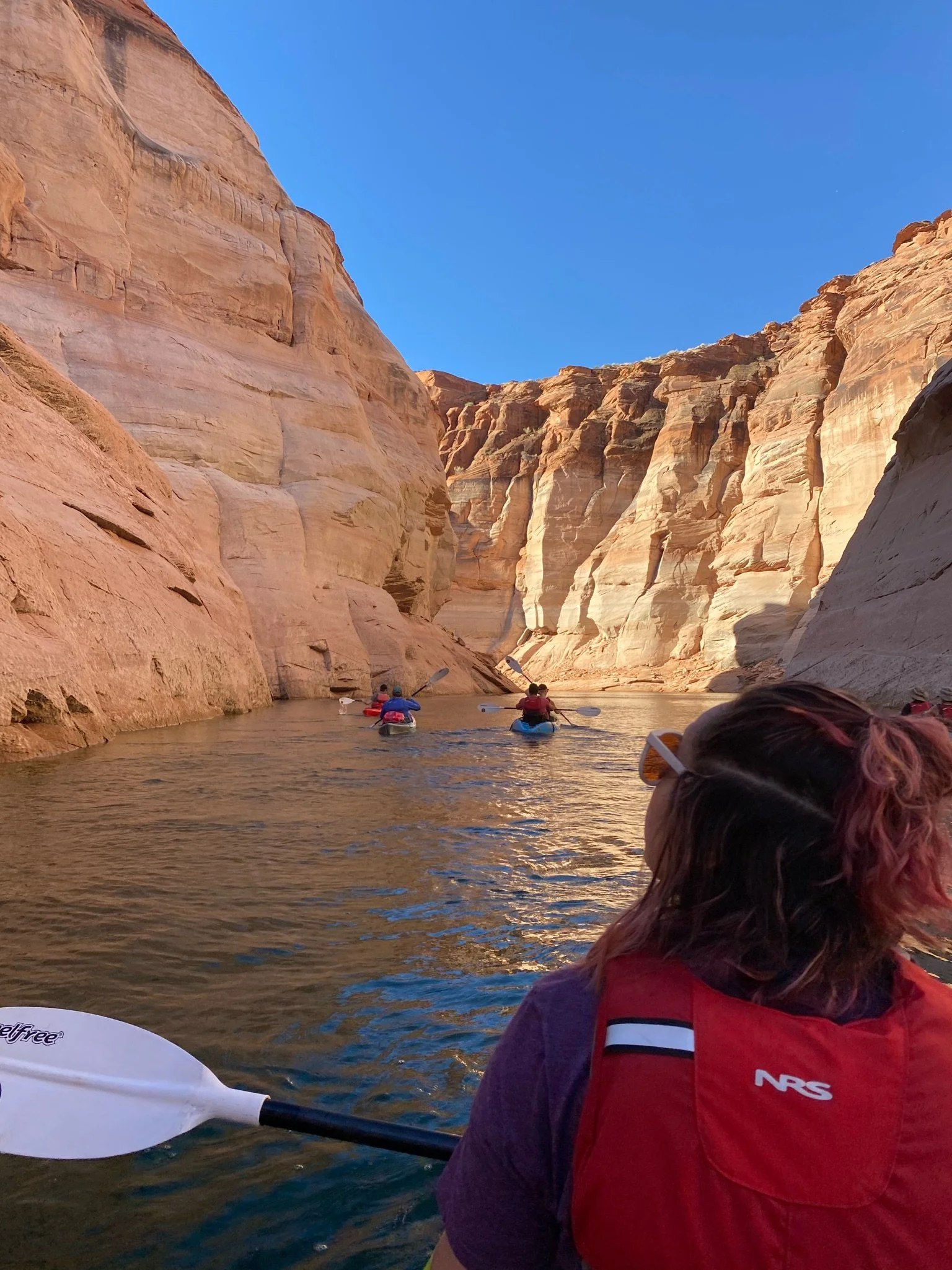 People kayaking through a narrow canyon with high red rock walls, blue sky above, during daytime.