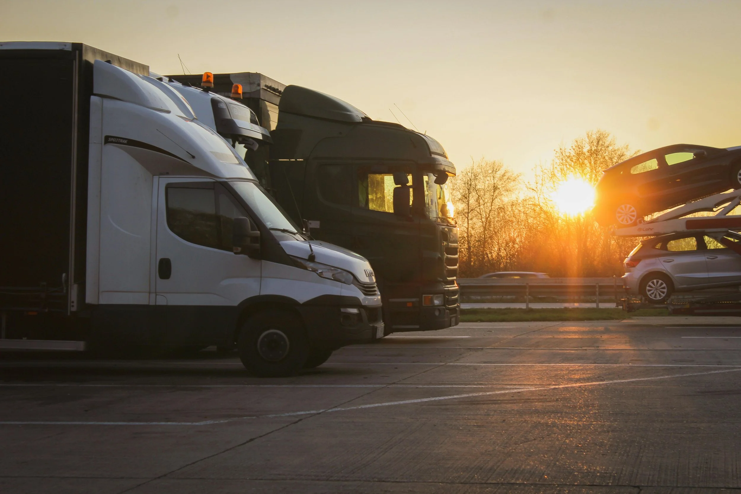 Vehicles, including trucks and cars, parked on a highway during sunset with trees in the background.