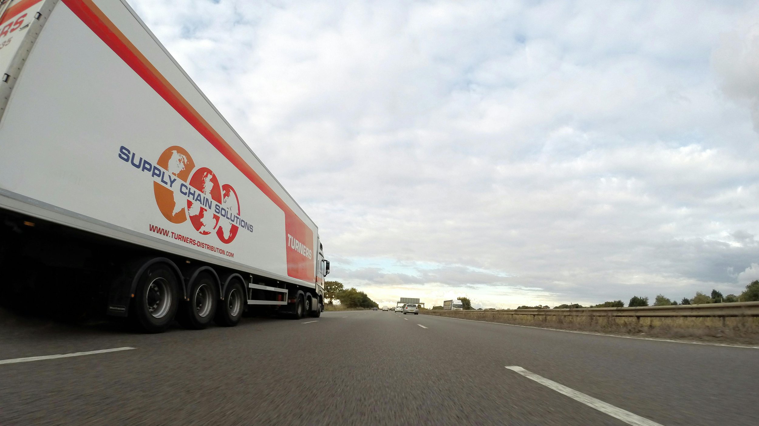 A highway with a large white truck labeled Supply Chain Solutions on the side, driving along a multi-lane road under a cloudy sky.