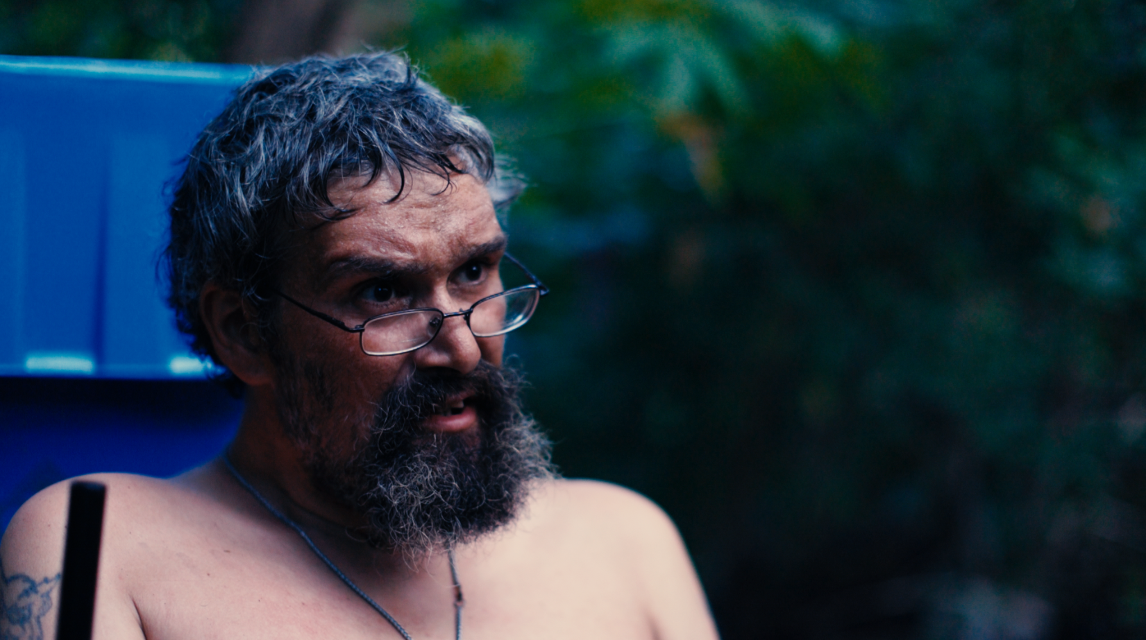 A shirtless man with glasses and a beard, outdoors, with a blue background and greenery.