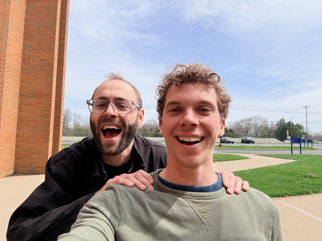 Two smiling young men taking a selfie outdoors on a cloudy day, standing in front of a brick building with a grassy area and parking lot in the background.