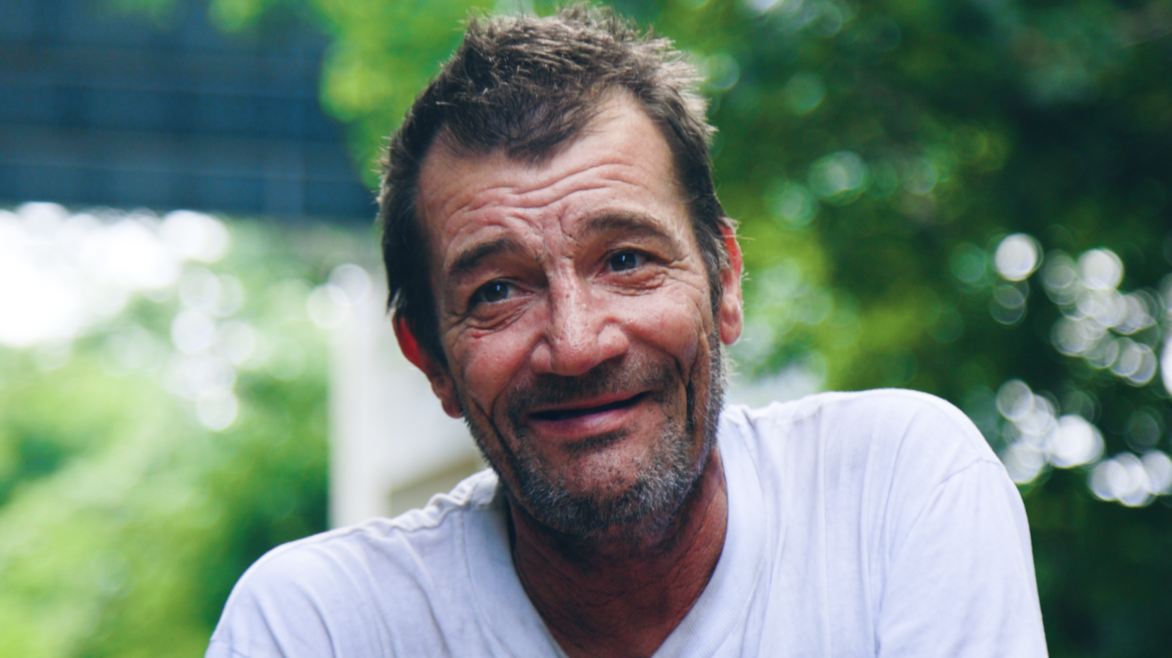 Smiling middle-aged man with dark hair, beard, and blue eyes outdoors, wearing a white shirt, with green trees in the blurry background.